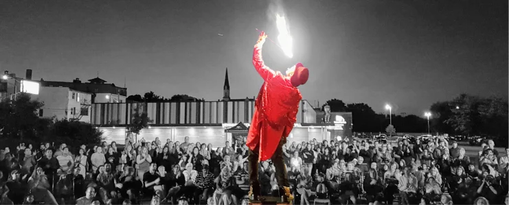 FireGuy is eating a large flame of fire while standing on a plank, on a pipe, on a box, in front of a crowd during a show.