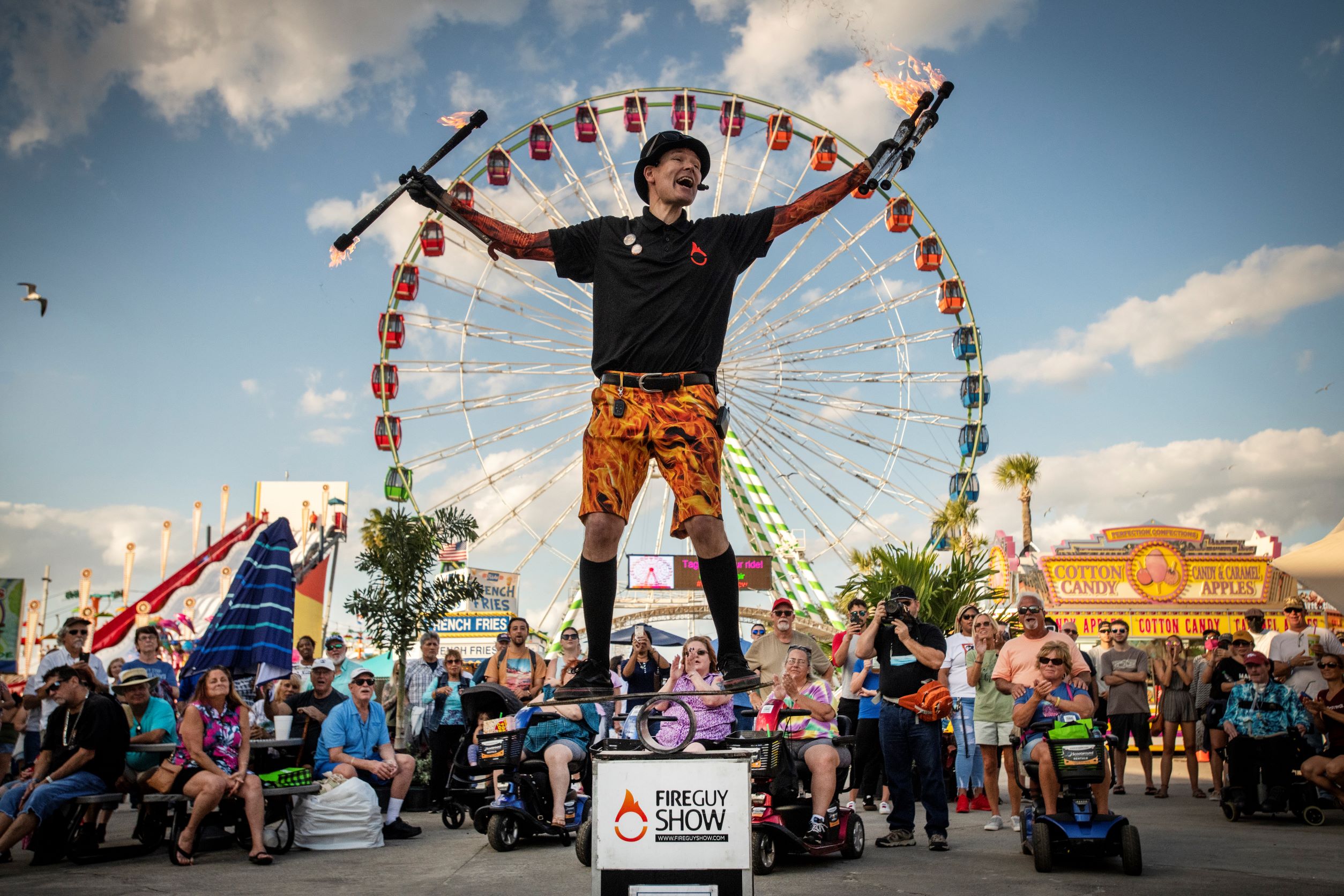 FireGuy standing on a board, on a pipe, on a box, talking to the crowd before or after a show while holding his fire sticks and baton.