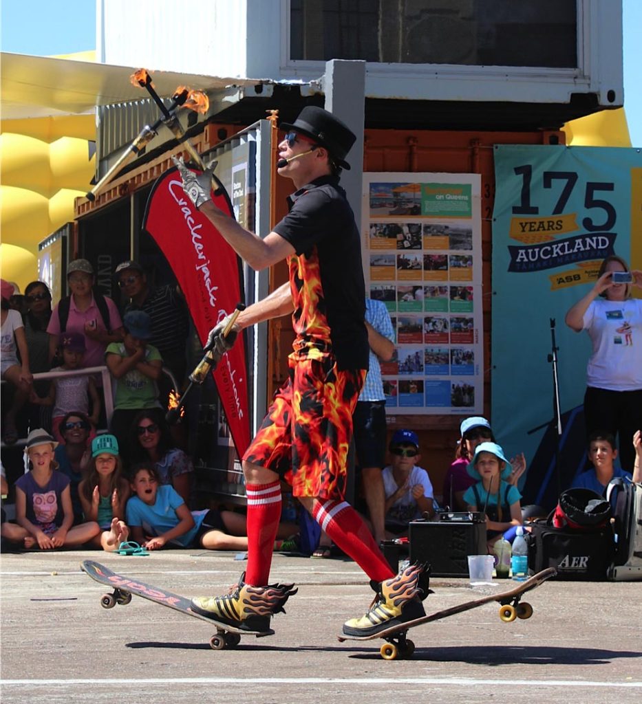 FireGuy is riding two skateboards, juggling three fire sticks, and talking to the crowd for a show in New Zealand.